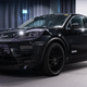 A 2024 Porsche Macan Electric prototype photographed inside a demonstration hall. the vehicle is painted black with vinyl appliques to obscure its design